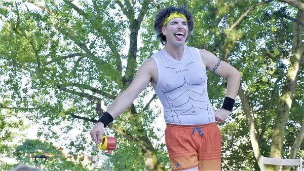 Un payaso con ropa deportiva, gorra y gafas, haciendo una pose cómica con una medalla gigante, en un escenario con elementos olímpicos.