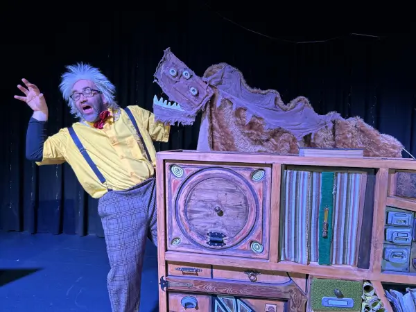 Un personaje disfrazado de monstrología interactúa con el público infantil en un escenario colorido, rodeado de elementos que evocan monstruos amigables.
