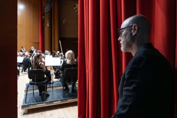 Un joven director de orquesta dirigiendo a la Real Filharmonía de Galicia en un escenario, con Baldur Brönnimann observando.