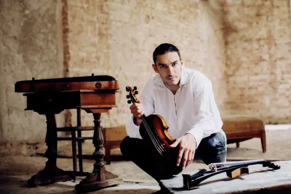 Sergey Malov tocando un violín o violoncello da spalla en el escenario, con una orquesta sinfónica de fondo.