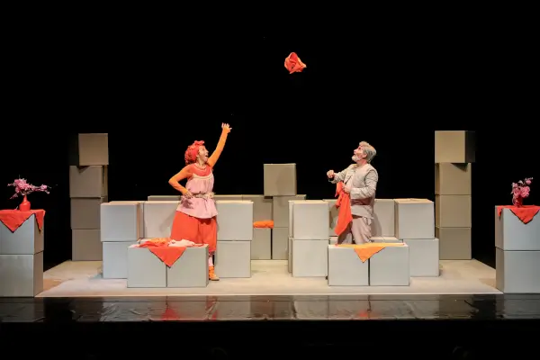 Escena de la obra 'Coralinda' con dos actores, uno vestido de gris y otro de colores, interactuando con cajas en un escenario minimalista.
