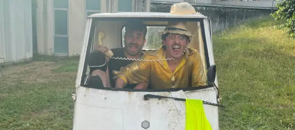 Dos cómicos ambulantes, Cosme y Damián, actúan en un escenario improvisado con un motocarro averiado de fondo.