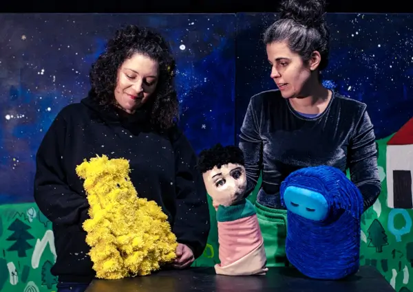 Un bebé en la cuna con dos figuras fantásticas, Meco y Sono, a su lado, en una obra de teatro de marionetas.