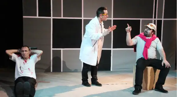 Tres actores en escena con vestuario colorido, interactuando con objetos que simulan productos de venta ambulante, en un ambiente teatral.