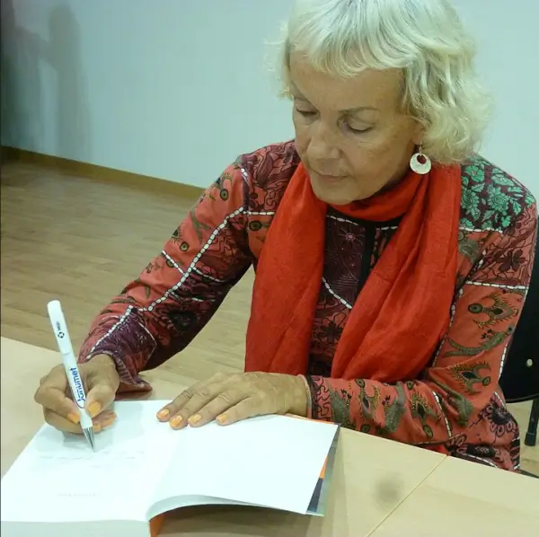 Retrato de la escritora y traductora checa Mónika Zgustová, sonriendo, con libros de fondo, en el contexto de una muestra bibliográfica.
